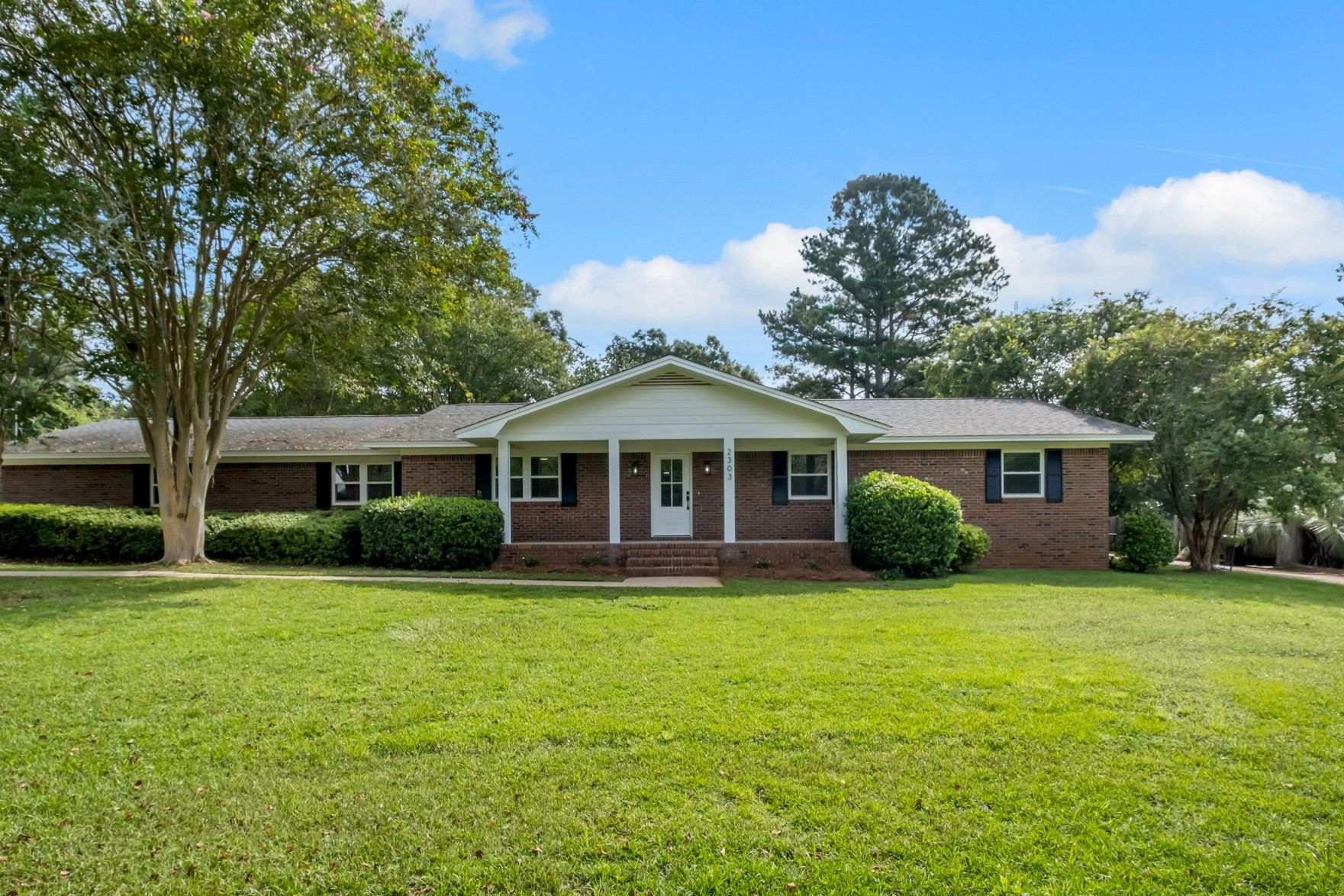 Front exterior: a long brick ranch with a white columned porch, set back behind a wide lawn and mature crepe myrtles